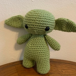 May include: A handmade, light green crochet doll with large, black eyes and oversized, pointed ears. The doll has a rounded head, small arms and legs, and is standing upright. The doll is made of a soft, textured yarn and is displayed on a wooden surface.