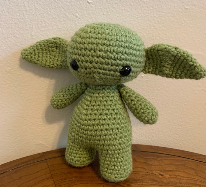 May include: A handmade, light green crochet doll with large, black eyes and oversized, pointed ears. The doll has a rounded head, small arms and legs, and is standing upright. The doll is made of a soft, textured yarn and is displayed on a wooden surface.