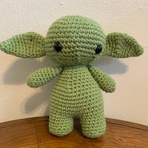 May include: A handmade, light green crochet doll with large ears, black eyes, and small arms and legs. The doll is standing upright and appears to be a character from a popular science fiction series. The doll is made of yarn.