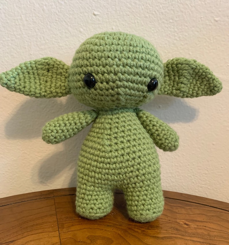 May include: A handmade, light green crochet doll with large ears, black eyes, and small arms and legs. The doll is standing upright and appears to be a character from a popular science fiction series. The doll is made of yarn.