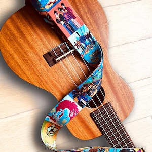 Könnte beinhalten: Eine Ukulele mit einem bunten Gitarrengurt mit Bildern der Beatles. Der Gurt hat ein mehrfarbiges Design mit Bildern der Bandmitglieder und Albumcovern.