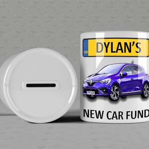 Könnte beinhalten: Weiße Keramik-Sparbüchse mit einem Schlitz oben. Die Vorderseite der Box zeigt ein blaues Auto mit einem Nummernschild, auf dem "DYLAN'S" steht, und dem Text "NEW CAR FUND".