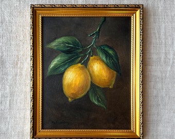 May include: A framed oil painting of two ripe lemons on a branch with green leaves. The lemons are yellow with textured surfaces. The painting is set against a dark background and framed in a gold ornate frame.