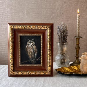 May include: A framed painting of an owl with brown and white feathers, perched on a branch. The owl has yellow eyes and is looking directly at the viewer. The painting is set against a dark background and is framed in a dark wood frame with gold accents.