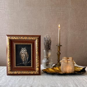 May include: A framed painting of an owl with brown and white feathers, perched on a branch. The frame is dark brown with gold accents. The painting is displayed on a table with a lit candle, a vase of dried lavender, and a gold leaf tray.