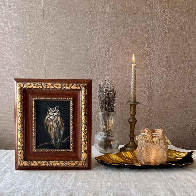 May include: A framed painting of an owl with brown and white feathers, perched on a branch. The frame is dark brown with gold accents. The painting is displayed on a table with a lit candle, a vase of dried lavender, and a gold leaf tray.