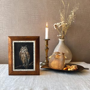 May include: A framed painting of an owl with brown and white feathers, perched on a branch. The painting is set against a dark background and is displayed on a table with a lit candle, a vase of dried flowers, and a burlap sack.