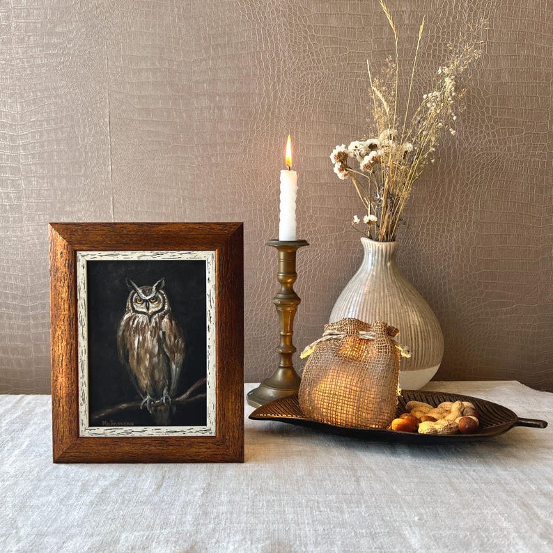 May include: A framed painting of an owl with brown and white feathers, perched on a branch. The painting is set against a dark background and is displayed on a table with a lit candle, a vase of dried flowers, and a burlap sack.