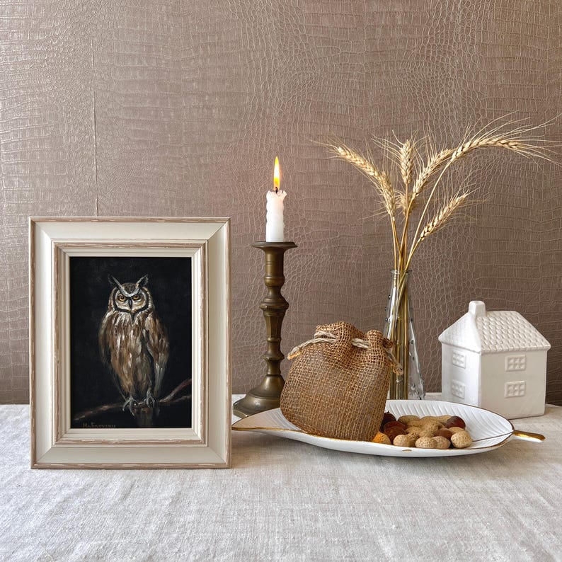 May include: A framed painting of an owl with brown and white feathers, set against a black background. The painting is displayed on a white table with a candle holder, a burlap sack, and a white ceramic house.