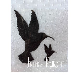 Peut inclure: Silhouette noire de deux colibris sur un fond blanc avec une texture scintillante. Le plus grand colibri vole les ailes déployées. Le plus petit colibri vole en dessous du plus grand. Le texte "French Kokotte" est imprimé sous les colibris.