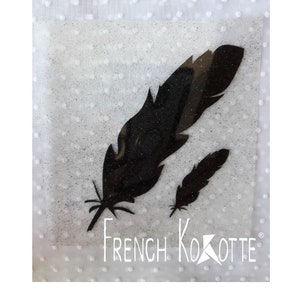 May include: Two black feather designs with glitter accents are displayed on a white background. The text "FRENCH KOKOTTE" is printed in white below the feathers. The feathers are different sizes.