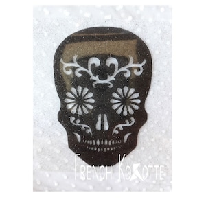 Peut inclure: Crâne de sucre noir et blanc avec des détails floraux. Le crâne est souligné de blanc et a des fleurs blanches et des volutes sur le front et les joues. L'arrière-plan est blanc avec une texture de paillettes subtile.