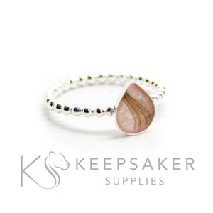 May include: A silver ring featuring a teardrop-shaped setting. The setting holds a pink resin with strands of hair. The ring band has a beaded design. The Keepsaker Supplies logo is in the lower left corner.