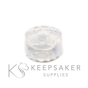 Pode incluir: Uma tarracha de brinco cilíndrica transparente com uma borda texturizada e estriada. A tarracha é feita de um material transparente e foi projetada para prender brincos. A imagem está em um fundo branco.