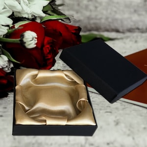 May include: A black gift box with a gold satin interior. The box is open and the satin lining is visible. The box is sitting on a white surface.