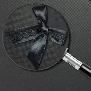 May include: A black satin ribbon with a lace trim tied into a bow. The bow is magnified by a magnifying glass.