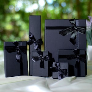 May include: Five black gift boxes of varying sizes with black satin ribbon bows. The boxes are arranged in a row on a white surface.