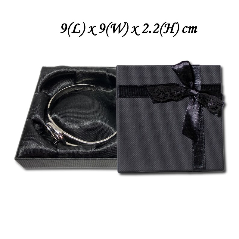 May include: A black gift box with a satin ribbon and bow. The box is 9 inches long, 9 inches wide, and 2.2 inches high. Inside the box is a black velvet-lined compartment with a silver bracelet.
