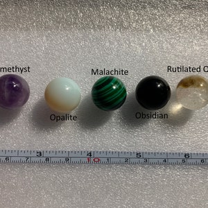 Puede incluir: Cinco esferas de piedras preciosas pulidas, cada una con un color y un patrón diferentes. De izquierda a derecha: amatista, opalita, malaquita, obsidiana y cuarzo rutilado.