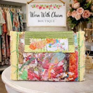 May include: A colorful, patchwork clutch with a mix of textures and patterns. The design incorporates green, pink, and yellow fabrics, with a floral accent. The clutch is rectangular with a fringed edge and the boutique name "Worn With Charm" is visible in the background.