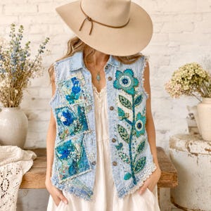 May include: A light blue denim vest featuring frayed armholes, a button-down front, and intricate floral embroidery in shades of blue and green. The vest is adorned with layered fabric patches and white lace trim along the bottom hem.