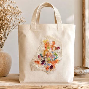 May include: A cream-colored canvas tote bag featuring a central decorative patch made of layered, frayed fabric scraps in vibrant shades of orange, yellow, green, and purple, accented with intricate embroidery and delicate white lace trim.