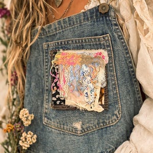 May include: Light blue denim overalls with a decorative patch on the pocket. The patch features a collage of fabrics in pink, white, and black. The overalls have a distressed look and metal hardware.