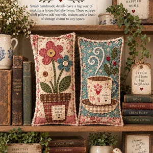 May include: Two rectangular fabric pillows with frayed edges. One features a potted flower design with the text "bloom small joys" and buttons. The other shows a teacup with steam and the text "a warmer day" on a tag, set on a saucer.