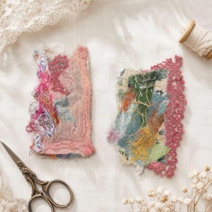 May include: Two textile art pieces with colorful, textured surfaces and pink lace edging. One piece is predominantly pink with blue and yellow accents, the other features green, blue, and yellow hues. A pair of scissors and a spool of lace are also present.