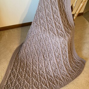 May include: A brown knitted throw blanket with a cable knit pattern. The blanket is draped over a wooden chair.