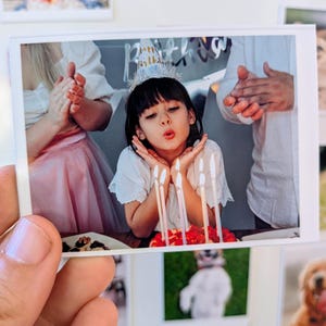 May include: A printed photograph of a child blowing out candles on a birthday cake. The child wears a party hat and is surrounded by clapping hands. Other photos are visible in the background.