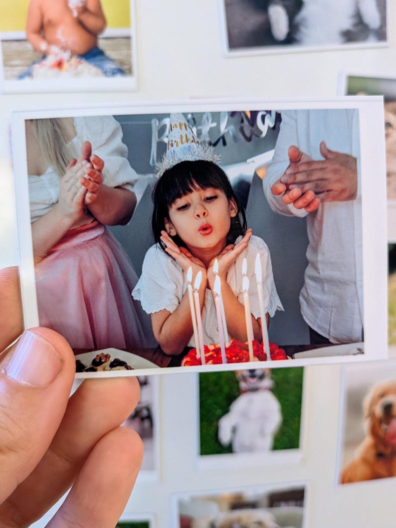 May include: A printed photograph of a child blowing out candles on a birthday cake. The child wears a party hat and is surrounded by clapping hands. Other photos are visible in the background.