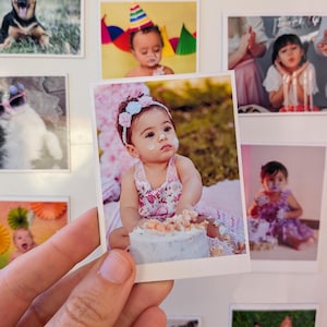 May include: A hand holds a square photo of a baby with a floral headband and dress, covered in frosting from a cake. Other photos of babies and pets are in the background, creating a collage.