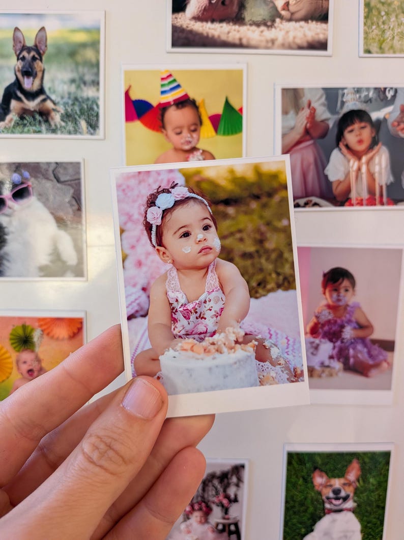May include: A hand holds a square photo of a baby with a floral headband and dress, covered in frosting from a cake. Other photos of babies and pets are in the background, creating a collage.
