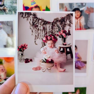 May include: A printed photograph of a baby wearing a pink tutu and a floral crown, sitting in front of a small cake on a decorative stand. The baby is surrounded by flowers and greenery, with other framed photos in the background.