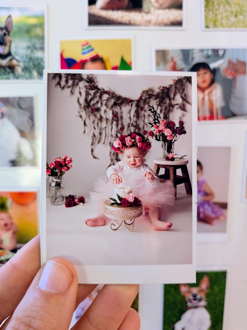 May include: A printed photograph of a baby wearing a pink tutu and a floral crown, sitting in front of a small cake on a decorative stand. The baby is surrounded by flowers and greenery, with other framed photos in the background.
