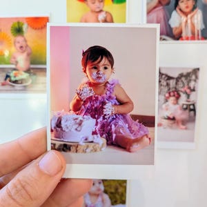 May include: A photograph of a baby girl covered in purple frosting, sitting in front of a cake. The girl is wearing a purple dress and has frosting on her face and hands. Other photos are visible in the background.
