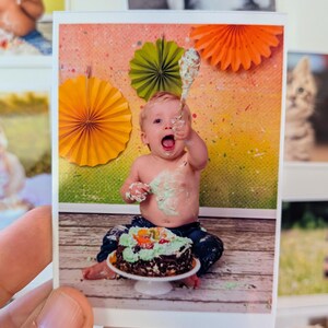 May include: A photograph of a baby celebrating a birthday. The baby is covered in frosting and holding a spoon. A chocolate cake with green frosting and fruit sits on a white cake stand. Colorful paper fans decorate the background.