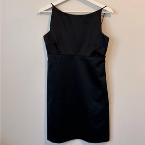 May include: A black, sleeveless dress with a square neckline and spaghetti straps. The dress is made of a shiny, satin-like material and hangs on a white hanger. The dress has a fitted bodice and a straight skirt, suitable for formal occasions.