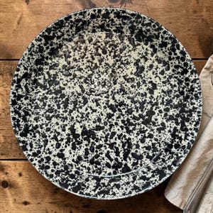 Può includere: Un piatto rotondo in ceramica con un motivo maculato nero e crema. Il piatto è su una superficie di legno, con un tovagliolo di lino e due forchette a destra. Il design del piatto ricorda un uovo maculato.