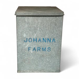 May include: A rectangular metal container with a lid. The container is grey with blue lettering that reads "JOHANNA FARMS." Faint text above the name reads "PROPERTY OF JOHANNA". The container has a slightly weathered appearance.