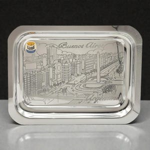 Puede incluir: Una bandeja rectangular plateada con esquinas redondeadas. La bandeja presenta un paisaje urbano grabado de Buenos Aires, Argentina. Las palabras "Buenos Aires" y "Argentina" están grabadas en la bandeja. La bandeja tiene un logotipo "LUBA" en la esquina superior izquierda.