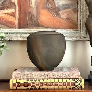 May include: A dark brown, matte-finished ceramic vessel with a rounded, bulbous body and a flat, circular top. The vessel features subtle, swirling color variations in shades of charcoal and bronze, resting on a stack of two hardcover books.