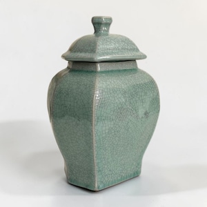 Peut inclure: Un pot en céramique vert clair avec un couvercle assorti, présentant une finition émaillée craquelée distincte. Le récipient a un corps de forme carrée aux coins arrondis et une petite poignée en forme de bouton sur le couvercle.