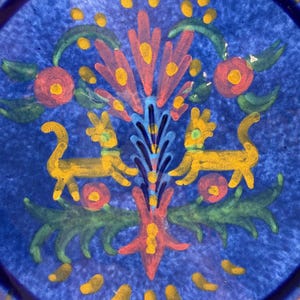 May include: A blue ceramic plate with a hand-painted design. The design features a central floral motif in red, yellow, and blue, flanked by two yellow animal figures. Green leaves and red flower accents complete the design. The plate's rim has a gold border.