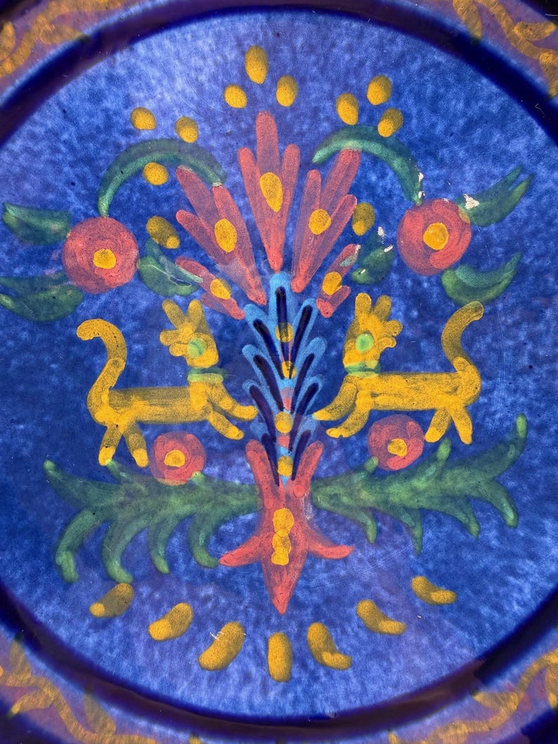 May include: A blue ceramic plate with a hand-painted design. The design features a central floral motif in red, yellow, and blue, flanked by two yellow animal figures. Green leaves and red flower accents complete the design. The plate's rim has a gold border.