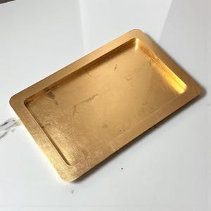 Może przedstawiać: Prostokątna taca w kolorze złotym z lekko podniesioną krawędzią. Taca ma wykończenie ze szczotkowanego metalu i jest umieszczona na białym tle. Taca jest pusta i wydaje się być wykonana z metalu.