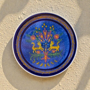 Puede incluir: Un plato redondo de cerámica con un centro azul que presenta un colorido diseño floral y animal. El plato tiene un borde blanco y un borde dorado. El diseño incluye elementos amarillos, rosas y verdes.