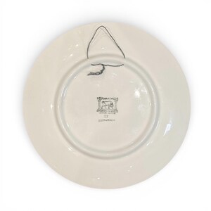 May include: A white ceramic plate with a black wire hanger. The back of the plate features a circular design with the text "KERAMIKOS" and other Greek text. The plate has a glossy finish.