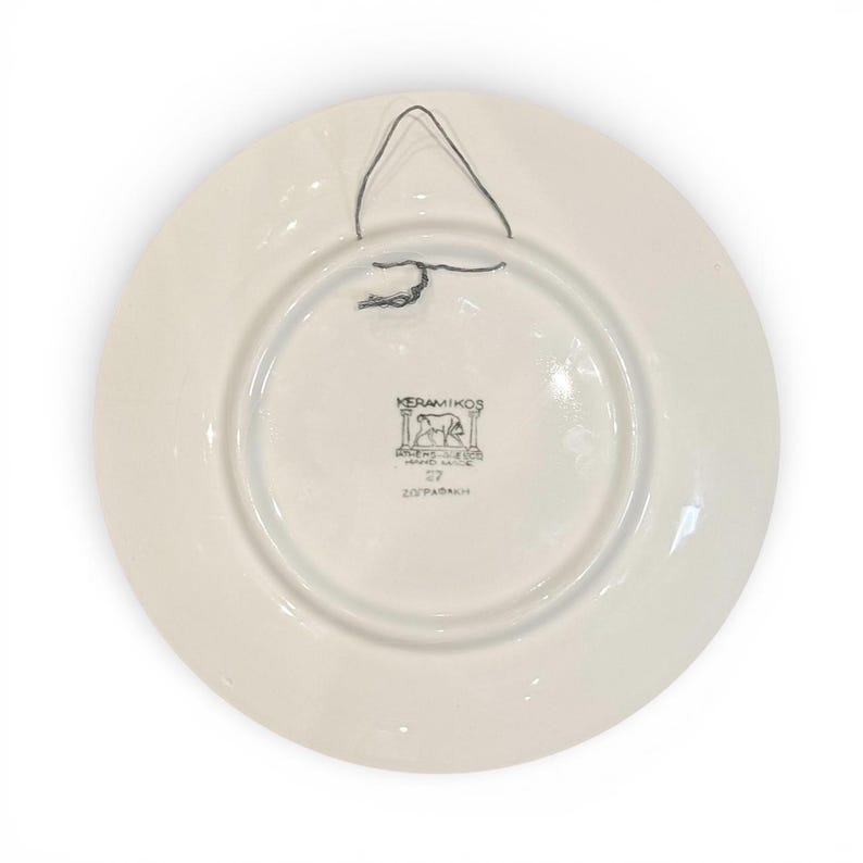 May include: A white ceramic plate with a black wire hanger. The back of the plate features a circular design with the text "KERAMIKOS" and other Greek text. The plate has a glossy finish.
